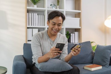 Genç bir adam kanepede rahat bir şekilde oturuyor, akıllı telefonunu kullanırken gülümsüyor ve bir kredi kartı tutuyor, çevrimiçi ödeme yapıyor. Sahne modern dijital alışveriş ve evdeki kolay ödemeyi yansıtıyor.