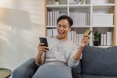 Genç bir adam kanepede rahat bir şekilde oturuyor, akıllı telefonunu kullanırken gülümsüyor ve bir kredi kartı tutuyor, çevrimiçi ödeme yapıyor. Sahne modern dijital alışveriş ve evdeki kolay ödemeyi yansıtıyor.