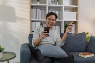 Genç bir adam kanepede rahat bir şekilde oturuyor, akıllı telefonunu kullanırken gülümsüyor ve bir kredi kartı tutuyor, çevrimiçi ödeme yapıyor. Sahne modern dijital alışveriş ve evdeki kolay ödemeyi yansıtıyor.