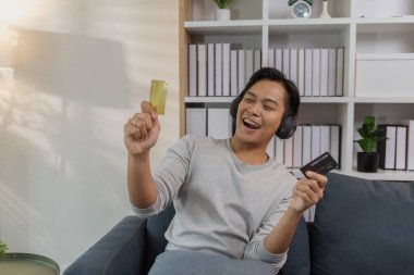 Genç bir adam kanepede rahat bir şekilde oturuyor, akıllı telefonunu kullanırken gülümsüyor ve bir kredi kartı tutuyor, çevrimiçi ödeme yapıyor. Sahne modern dijital alışveriş ve evdeki kolay ödemeyi yansıtıyor.