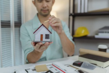 Bir adam tek eliyle küçük bir ev modelini tutarken bir yandan da çatıyı kalemle işaret ediyor. Sahne, modern bir çalışma alanında mimari tasarım, planlama veya gayrimenkul sunumu taşır..