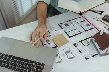 Defter ve dizüstü bilgisayardan bahsederken kalem kullanarak ev planı üzerinde çalışan bir mimar. Masanın üzerine küçük bir ev modeli ve zemin planı yerleştiriliyor. Detaylı mimari düzenini gösteriyor..