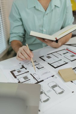 Elinde bir defter ve kalem tutarken evin planını gözden geçiren mimar. Çalışma alanı, temiz bir ortamda dizayn ve tasarım planlamasına odaklanan bir dizüstü bilgisayar ve mimari çizimler içerir..