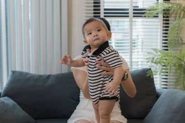 Genç bir anne, bebeğini sevgiyle kucaklar ve bir kanepede emzirir. Erken çocukluk ve aile hayatının anne bakımını, bağlarını ve besleyici anlarını sembolize eder..