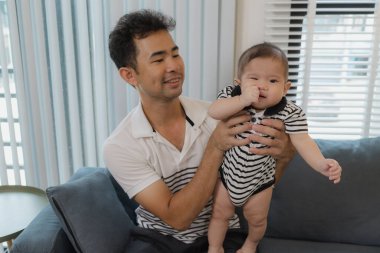 Neşeli bir baba bebeğini kollarında tutuyor, evdeki kanepede oturuyor. Bebek meraklı ve sakin görünürken baba sıcak bir gülümsemeyle aile bağlarını yakalıyor..