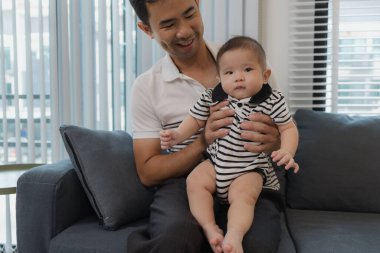 Neşeli bir baba bebeğini kollarında tutuyor, evdeki kanepede oturuyor. Bebek meraklı ve sakin görünürken baba sıcak bir gülümsemeyle aile bağlarını yakalıyor..