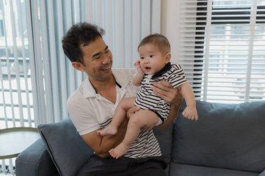 Neşeli bir baba bebeğini kollarında tutuyor, evdeki kanepede oturuyor. Bebek meraklı ve sakin görünürken baba sıcak bir gülümsemeyle aile bağlarını yakalıyor..