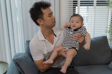 Neşeli bir baba bebeğini kollarında tutuyor, evdeki kanepede oturuyor. Bebek meraklı ve sakin görünürken baba sıcak bir gülümsemeyle aile bağlarını yakalıyor..