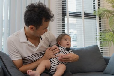 Neşeli bir baba bebeğini kollarında tutuyor, evdeki kanepede oturuyor. Bebek meraklı ve sakin görünürken baba sıcak bir gülümsemeyle aile bağlarını yakalıyor..