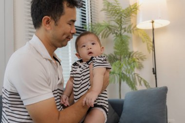 Neşeli bir baba bebeğini kollarında tutuyor, evdeki kanepede oturuyor. Bebek meraklı ve sakin görünürken baba sıcak bir gülümsemeyle aile bağlarını yakalıyor..