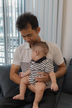 Neşeli bir baba bebeğini kollarında tutuyor, evdeki kanepede oturuyor. Bebek meraklı ve sakin görünürken baba sıcak bir gülümsemeyle aile bağlarını yakalıyor..