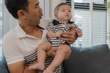 Neşeli bir baba, bebeğiyle oturma odasında bağ kuruyor. Bebek ellerini tutarken sıcak bir şekilde gülümsüyor. Sevgi, neşe ve ebeveynliğin mutluluğunu yansıtan an..