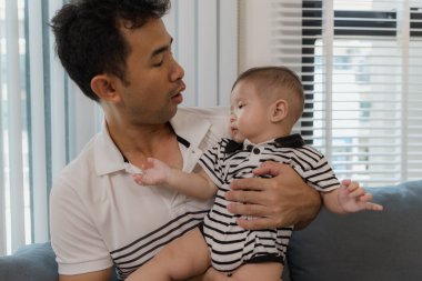 Neşeli bir baba, bebeğiyle oturma odasında bağ kuruyor. Bebek ellerini tutarken sıcak bir şekilde gülümsüyor. Sevgi, neşe ve ebeveynliğin mutluluğunu yansıtan an..