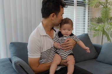 Neşeli bir baba, bebeğiyle oturma odasında bağ kuruyor. Bebek ellerini tutarken sıcak bir şekilde gülümsüyor. Sevgi, neşe ve ebeveynliğin mutluluğunu yansıtan an..