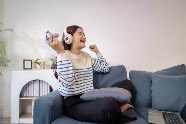 Genç bir kadın, elinde oyun kumandası ve dizüstü bilgisayarıyla video oyunu oynarken kulaklık takarak koltukta oturuyor. Neşeli ifadesi neşe ve heyecan gösteriyor..