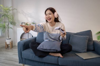 Genç bir kadın, elinde oyun kumandası ve dizüstü bilgisayarıyla video oyunu oynarken kulaklık takarak koltukta oturuyor. Neşeli ifadesi neşe ve heyecan gösteriyor..