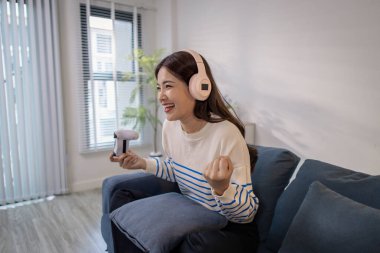 Genç bir kadın, elinde oyun kumandası ve dizüstü bilgisayarıyla video oyunu oynarken kulaklık takarak koltukta oturuyor. Neşeli ifadesi neşe ve heyecan gösteriyor..