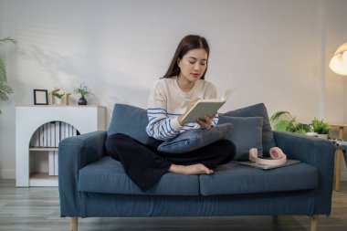 Genç bir kadın kanepede rahatça oturuyor, defterine odaklanarak yazarken gülümsüyor. Kulaklıkları ve dizüstü bilgisayarı yakınlarda dinleniyor, rahat bir çalışma ortamı yaratıyor..
