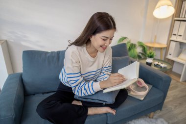 Genç bir kadın kanepede rahatça oturuyor, defterine odaklanarak yazarken gülümsüyor. Kulaklıkları ve dizüstü bilgisayarı yakınlarda dinleniyor, rahat bir çalışma ortamı yaratıyor..