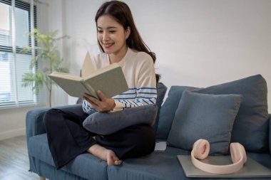 Genç bir kadın kanepede rahatça oturuyor, defterine odaklanarak yazarken gülümsüyor. Kulaklıkları ve dizüstü bilgisayarı yakınlarda dinleniyor, rahat bir çalışma ortamı yaratıyor..