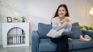 Genç bir kadın kanepede rahatça oturuyor, defterine odaklanarak yazarken gülümsüyor. Kulaklıkları ve dizüstü bilgisayarı yakınlarda dinleniyor, rahat bir çalışma ortamı yaratıyor..