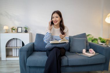 Genç bir kadın kanepede rahat bir şekilde oturur, elinde bir defter tutar ve akıllı telefonuyla gülerek konuşur. Rahatlamış görünüyor, notları okumak ve sohbet etmek arasında birden fazla iş yapıyor..