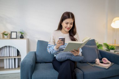 Genç bir kadın kanepede rahatça oturur, elinde kahve fincanı tutarken defter okur. Rahatlamış ve odaklanmış görünüyor. Evde sakin bir çalışma ya da planlama anının tadını çıkarıyor..