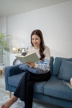 Genç bir kadın kanepede rahatça oturur, elinde kahve fincanı tutarken defter okur. Rahatlamış ve odaklanmış görünüyor. Evde sakin bir çalışma ya da planlama anının tadını çıkarıyor..