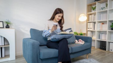 Genç bir kadın kanepede rahatça oturur, elinde kahve fincanı tutarken defter okur. Rahatlamış ve odaklanmış görünüyor. Evde sakin bir çalışma ya da planlama anının tadını çıkarıyor..
