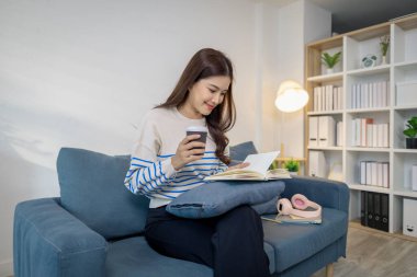 Genç bir kadın kanepede rahatça oturur, elinde kahve fincanı tutarken defter okur. Rahatlamış ve odaklanmış görünüyor. Evde sakin bir çalışma ya da planlama anının tadını çıkarıyor..