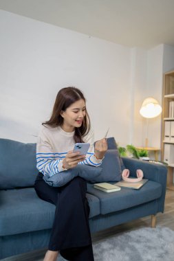 Bir elinde akıllı telefon, diğer elinde kredi kartı, dijital finans ve e-ticaret yaşam tarzını temsil eden bir genç kadın, evinde bir kanepede oturmuş gülümsüyor..