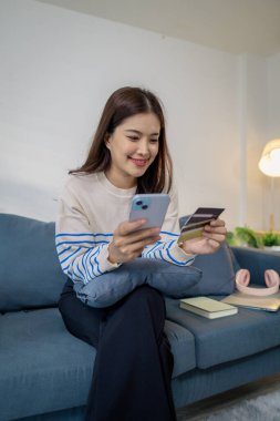 Bir elinde akıllı telefon, diğer elinde kredi kartı, dijital finans ve e-ticaret yaşam tarzını temsil eden bir genç kadın, evinde bir kanepede oturmuş gülümsüyor..