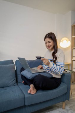 Genç bir kadın, elinde kredi kartı tutarken bir dizüstü bilgisayar kullanarak kanepeye oturuyor, güvenli online ödemeyi ve modern yaşam tarzını temsil ediyor. Evden alışveriş yaparken kendine güvenerek gülümsüyor..