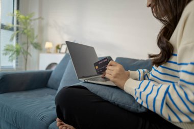 Genç bir kadın, elinde kredi kartı tutarken bir dizüstü bilgisayar kullanarak kanepeye oturuyor, güvenli online ödemeyi ve modern yaşam tarzını temsil ediyor. Evden alışveriş yaparken kendine güvenerek gülümsüyor..