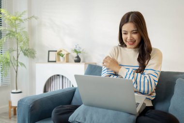 Genç bir kadın kanepede rahat bir şekilde oturuyor, dizüstü bilgisayar kullanıyor, odaklanarak ve sevinçle gülümsüyor. İşiyle ya da internet iletişimiyle meşgul görünüyor, sıcak bir yaşam tarzı konsepti yaratıyor..