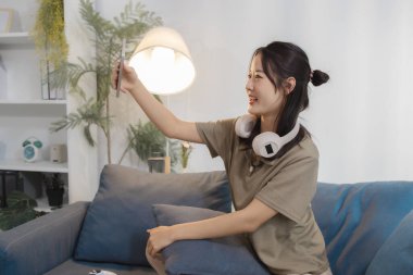 Kanepede oturan genç bir kadın, kulaklıkları boynunda, akıllı telefonunu tutarken gülümsüyor, rahat ve rahat bir evde selfie çekiyor gibi görünüyor..