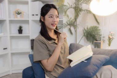 Genç bir kadın, modern bir ofisteki koltukta rahat bir şekilde oturur elinde bir defter tutarak düşünceli bir şekilde yazar. Odaklanmış ve rahat görünüyor, sakin bir kişisel yansımanın tadını çıkarıyor..