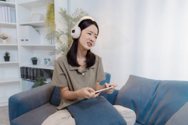 Kanepede oturan ve kulaklık takan genç bir kadın, elinde akıllı bir telefon tutarken hayal kırıklığı ya da hayal kırıklığı yaşarken evde cep telefonu oyunu oynarken hayal kırıklığına uğramış görünüyor..
