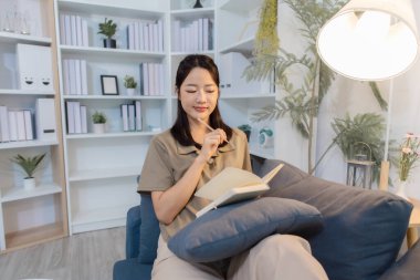 Genç bir kadın, modern bir ofisteki koltukta rahat bir şekilde oturur elinde bir defter tutarak düşünceli bir şekilde yazar. Odaklanmış ve rahat görünüyor, sakin bir kişisel yansımanın tadını çıkarıyor..