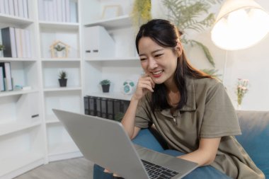 Genç bir kadın elinde dizüstü bilgisayarla kanepede oturuyor. Ekrana bakıp gülümsüyor. Rahatlamış ve meşgul görünüyor, çevrimiçi iletişimden, uzaktan çalışmadan ya da evde dijital öğrenmeden hoşlanıyor..