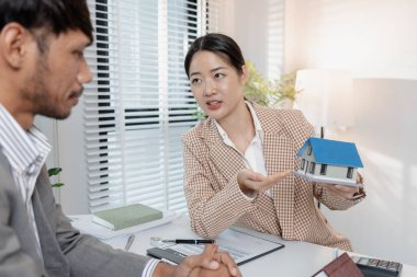 Bir emlakçı, şartları açıklamak için ev modelleri ve hesap makinesi kullanarak bir müşteriyle emlak sözleşmelerini tartışır. Sahne, müzakere, karar verme ve mali planlamayı vurguluyor.