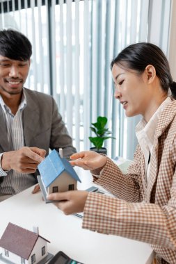 Bir emlakçı, şartları açıklamak için ev modelleri ve hesap makinesi kullanarak bir müşteriyle emlak sözleşmelerini tartışır. Sahne, müzakere, karar verme ve mali planlamayı vurguluyor.