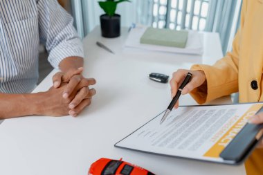 Sarı ceketli bir kadın satıcının, erkek bir müşteriye araba modellerini göstermesi ve anahtarları teslim etmesi ile ilgili detayları açıkladığı bir ofiste bir araba satışı pazarlığı gerçekleşiyor..