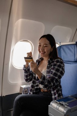 Gülümseyen Asyalı kadın bir kredi kartı tutarken ve uçakta akıllı telefon kullanırken, başarılı bir online ödeme veya uçuş sırasında alışveriş yaptıktan sonra heyecanını gösteriyor..