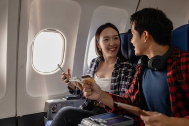 Uçakta birlikte oturan gülümseyen Asyalı çift ellerinde nakit para, kredi kartı ve pasaport tutuyorlar. Yolculuğa hazır olduklarını ve heyecanlandıklarını gösteriyorlar. Uçuş sırasında mutlu ve rahat görünüyorlar..