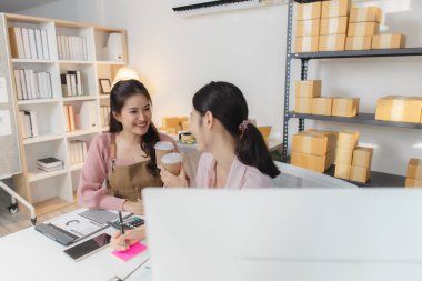 İki genç kadın küçük bir iş ofisinde birlikte çalışır, paketleri düzenler, mali raporları kontrol eder ve dijital cihazlar üzerindeki siparişleri yönetir, takım çalışmasını ve girişimciliği temsil eder..