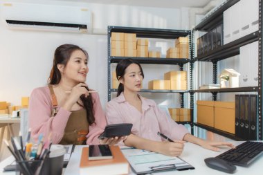 İki genç kadın küçük bir iş ofisinde birlikte çalışır, paketleri düzenler, mali raporları kontrol eder ve dijital cihazlar üzerindeki siparişleri yönetir, takım çalışmasını ve girişimciliği temsil eder..