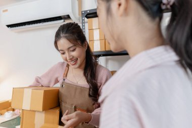 İki genç kadın, küçük bir işletme ofisinde birlikte çalışıyorlar, etrafları paket ve belgelerle çevrili. Görevleri tartışıyor, envanterleri kontrol ediyor ve takım çalışması ve odaklanmayla paketleri hazırlıyorlar..