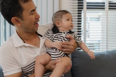 Neşeli bir baba, bebeğiyle oturma odasında bağ kuruyor. Bebek ellerini tutarken sıcak bir şekilde gülümsüyor. Sevgi, neşe ve ebeveynliğin mutluluğunu yansıtan an..