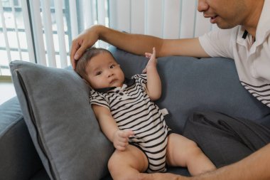 Neşeli bir baba, bebeğiyle oturma odasında bağ kuruyor. Bebek ellerini tutarken sıcak bir şekilde gülümsüyor. Sevgi, neşe ve ebeveynliğin mutluluğunu yansıtan an..